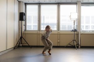Tanz-Performerin in geduckter Haltung, eine Hand zur Betrachter:in ausstreckend; im Hintergrund stehen Ton- und Lichttechnik